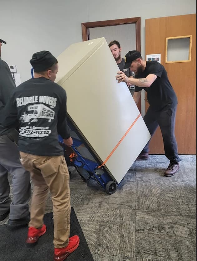 Reliable Movers crew carefully moving a heavy loaded fireproof filing cabinet inside an office building using professional moving equipment