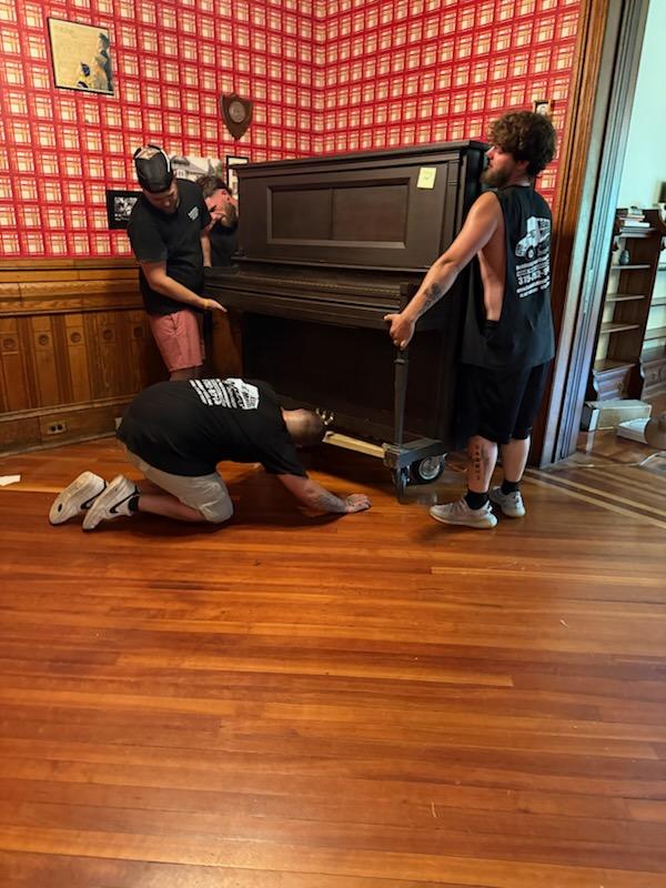 Reliable Movers carefully moving a heavy upright piano inside a historic Victorian home in Clinton, New York.