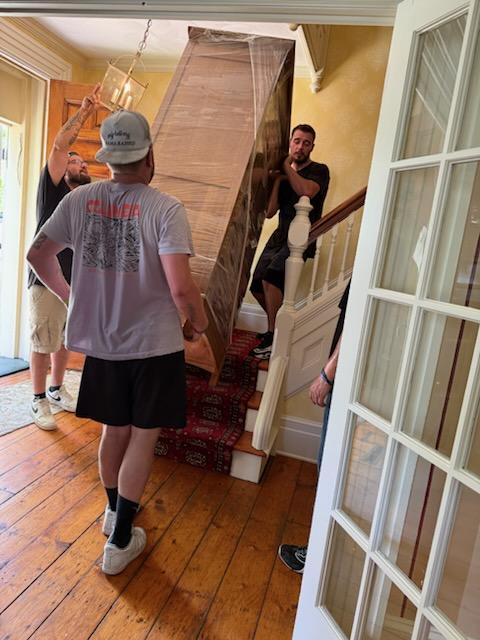 Reliable Movers carefully moving a large wrapped cabinet down a staircase inside a Victorian home in Clinton, New York.